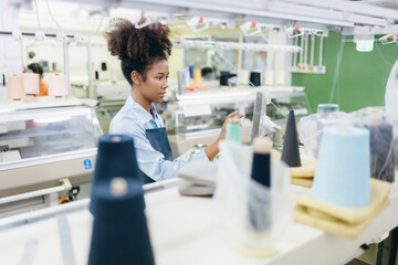 American female designer as seamstress uses circuit board to order clothes of sewing machine in industrial factory, weaving business holding laptop. Waiting to export products to the world market