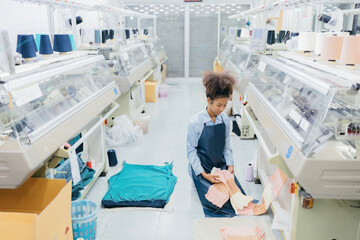 American female designer working as a seamstress sits and weaves fabric with thread next to a...