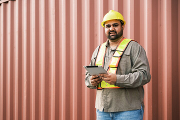An attractive Indian male industrial engineer in yellow hard hat, safety vest with a blurred container yard in background. Working in the logistics center. Inspector, supervisor in container terminal. - 756226159