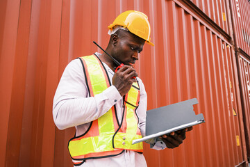 Portrait of an African man engineer or industrial worker in white hard hat, high-visibility Vest, talking on walkie-talkie radio at container yard. Inspector or safety supervisor in container terminal - 756226332