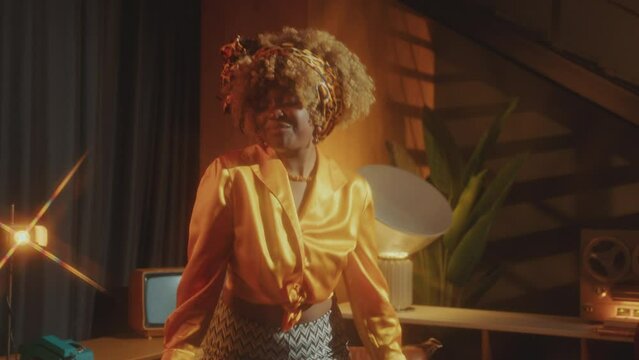 Young pretty African American woman in stylish outfit having fun and dancing to disco music in studio with retro interior and warm light
