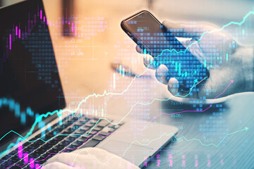 Close up of businessman hands using laptop and smartphone at desk with creative glowing candlestick forex chart on blurry background. Financial trade and market concept. Double exposure.