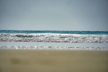 A sandy beach with water foaming around it and waves coming in from the ocean. Blurred brown sand on the beach.Blue water in the ocean. Arabian sea water shining in sunlight on sunny day in Goa,India.