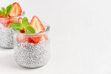 Homemade healthy natural chia pudding made with seeds soaked in vegetarian plant based milk decorated with sliced juicy strawberries topping and mint leaf served on white wooden table with copy space