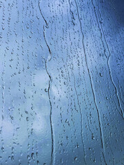 Rain drops sliding down a window glass, grey-blue cloudy sky blurred on the background. Rainy weather concept. Close-up shot