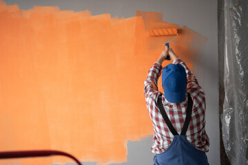 Rear view of painter man painting the wall with paint roller and orange color. Big empty space.