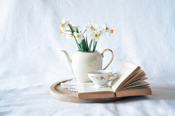 Still life with a blooming bouquet of white daffodils in an elegant porcelain teapot, a baroque porcelain coffee cup and an old book on a textured white background