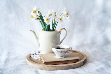 Still life with a blooming bouquet of white daffodils in an elegant porcelain teapot, a baroque porcelain coffee cup and an old book on a textured white background