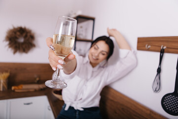 Young tipsy woman hold drink glass of champagne alcohol in kitchen sitting alone chilling relaxing having fun dancing on worktop wooden counter. Celebrating holiday, festive, celebration