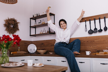 Young tipsy woman hold drink glass of champagne alcohol in kitchen sitting alone chilling relaxing having fun dancing on worktop wooden counter. Celebrating holiday, festive, celebration
