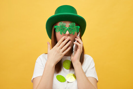 Fun and festive Irish party. Surprised Caucasian woman wearing green leprechaun hat and clover glasses standing isolated over yellow background having phone conversation hearing something shocked - Powered by Adobe