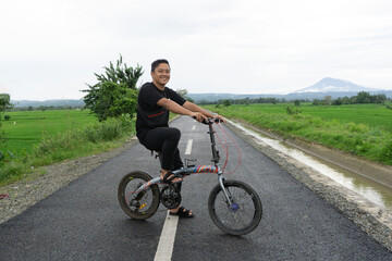 Fototapeta premium Happy Asian man riding a bicycle at the morning on the asphalt road. Cycling with mountain and paddy rice field view at the background. 