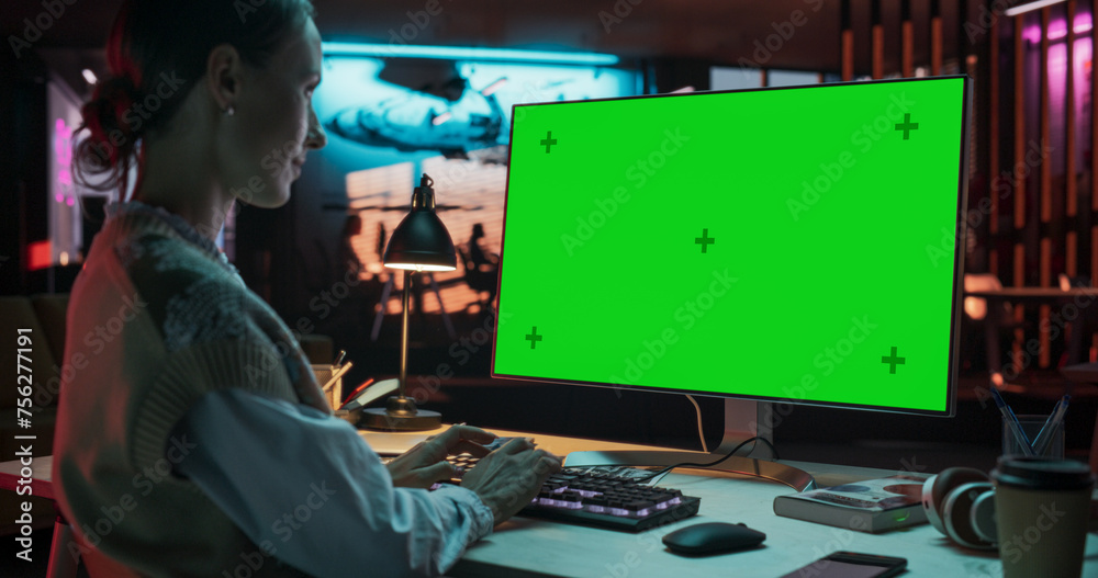 Aufkleber over the shoulder: young woman sitting at her desk using desktop computer with mock-up green screen.