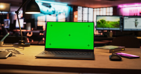 Mock-up Green Screen on a Laptop Standing on the top of a Wooden Table. In the Background Stylish Modern Office Studio in the Evening with Neon Lights. Desk of Creative Designer 