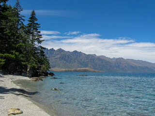 Gravel beach on beautiful, pristine Lake Wakatipu, surrounded by tall green pine trees and with a view of the Remarkables mountain range at Queenstown on New Zealand's South Island