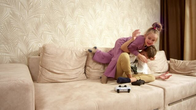 Two happy emotional children brother and sister playing game console using joystick or controller while sitting on the sofa at home. The concept of family vacation, entertainment and video games.