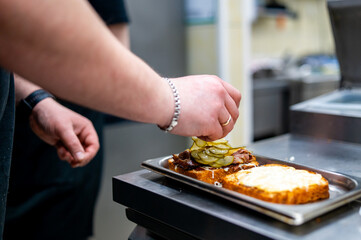 Obraz na płótnie Canvas chef adding sliced pickles to a grilled sandwich in a professional kitchen. The sandwich consists of two slices of toasted bread—one topped with grilled meat and the other with a spread.