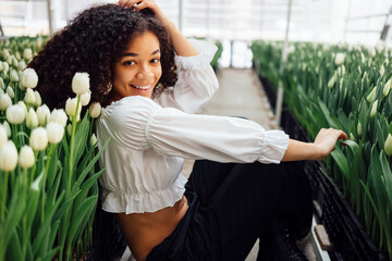 Female portrait of attractive darkskinned girl sitting on path among tulips in greenhouse