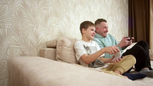 A father spends time with his little son, sitting on the couch and playing a game console. Weekend activities, free time, home entertainment and video games concept.