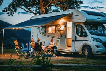 A family spends quality time together outside their RV at a campsite during a calm evening under the twilight sky - obrazy, fototapety, plakaty