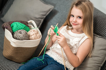 Cute blond little girl knitting on sofa at home enjoying leisure - obrazy, fototapety, plakaty