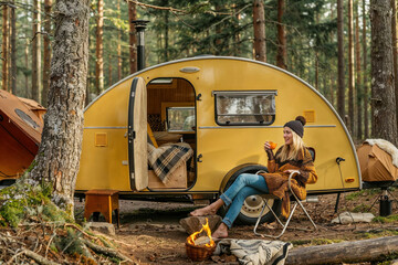 In a peaceful woodland setting, a woman enjoys a hot drink beside a classic yellow camper with a campfire nearby