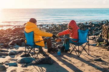 Two people on beach chairs together on winter beach, making hot tea, coffee on sunny blue sky day and the sea in background. British cold winter. Lifestyle tourism concept - obrazy, fototapety, plakaty