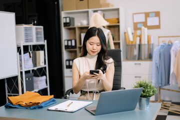  Asian young woman on desk in office of fashion designer and holds tablet, laptop and smartphone on...