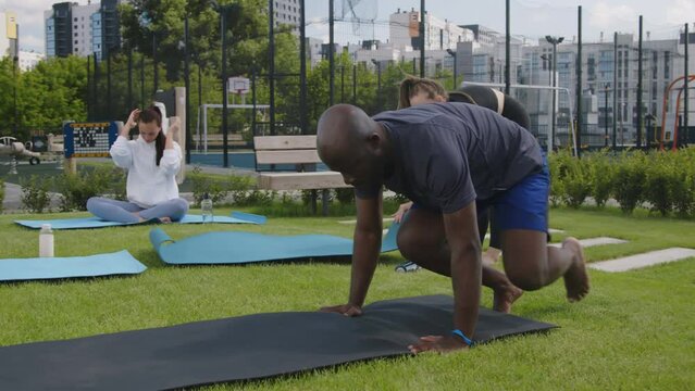 Side full footage of young African American man in activewear spreading mat on lawn in rooftop garden before having yoga practice in morning with female neighbors outdoors