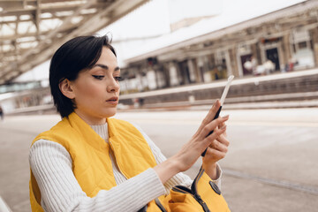 young brunet woman  in yellow jacket holding passport and tickets, and waiting for train at railway...