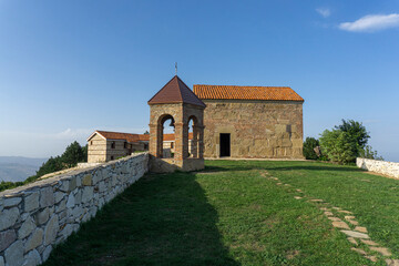 Fototapeta na wymiar St. Giorgi church of Udzo monastery and bell tower. Tile orange roof, brick and stone wall. Brick arches of the bell tower. Grass yard, blue clear sky.
