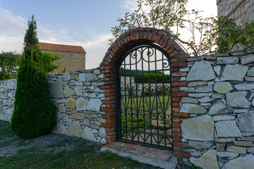 Beautiful iron gate with patterns in a stone wall with a brick arch. A church with a tiled roof is visible. Blue sky.