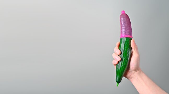 Unrecognizable person holding a cucumber with Red condom. Panoramic image with copy space. Safe sex concept.	