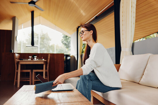 Relaxed Woman Smiling and Working on Laptop in Home Office An inspiring image capturing a young woman, comfortably seated on a sofa in her cozy living room She sports stylish glasses and a cheerful