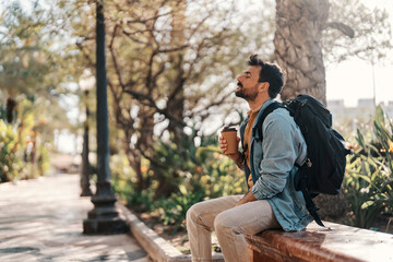 Portrait of a trendy man with backpack sitting on a city street and taking a break with his coffee to go.