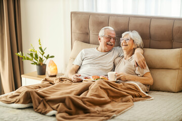 A happy senior couple is having breakfast in bed and hugging.