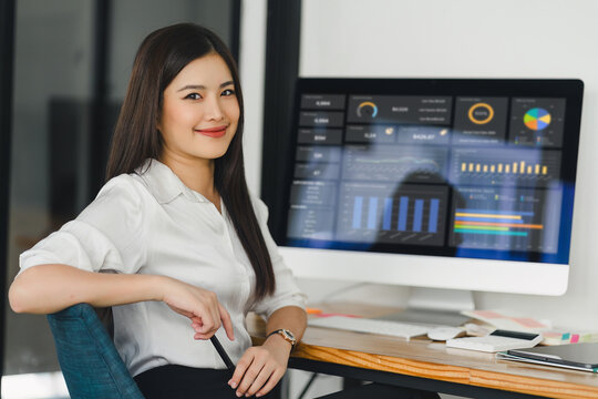 Asian businesswoman or accountant are analyzing data charts, graphs, and a dashboard on desktop screen in order to prepare a statistical report and discuss financial data in an office.