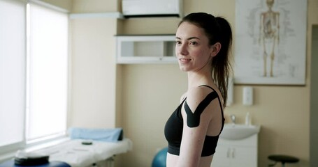 Glad woman with kinesiology tape. Positive female with kenesiology tape on shoulder smiling and looking at camera in daytime in rehabilitation clinic.