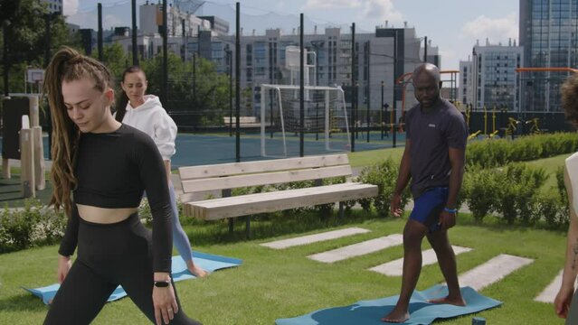 Arc footage of group of diverse women and man practicing yoga on mats in condo park and performing virabhadrasana or warrior pose