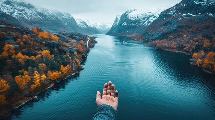  First-person perspective of a hand reaching out over a fjord surrounded by autumnal forests and snow-capped mountains. - obrazy, fototapety, plakaty