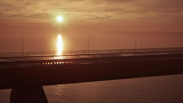 The sun is seen lowering in the sky as it sets over the ocean, with a bridge prominently featured in the foreground.