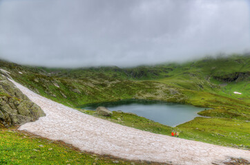 Mountain valley with a lake against a background of fog. Small figures of tourists in the distance.