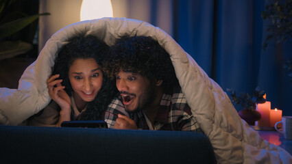 Multiracial couple man and woman at night home win mobile phone bet good news achievement triumph excited with online winning victory smartphone high five at evening couch under blanket gadget addict