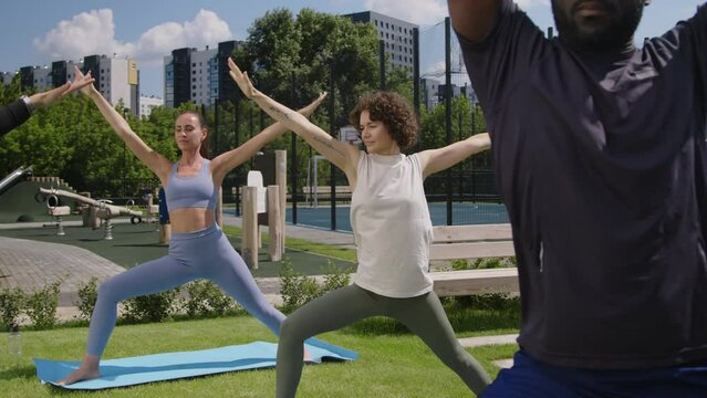 Side footage of group of diverse people practicing yoga outdoors in condo park and doing warrior pose or virabhadrasana