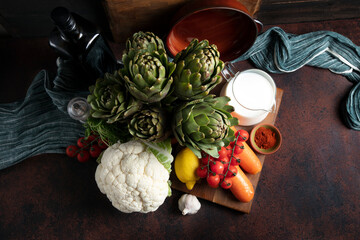 Dark-toned still life featuring artichokes, cauliflower, lemon, carrot, garlic, cherry tomatoes,...