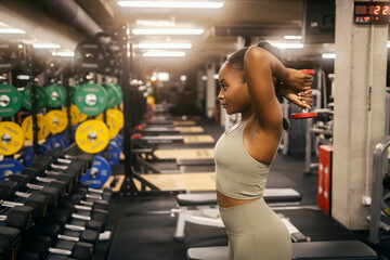Side view of a fit black sportswoman doing overhead triceps extension with dumbbell at the gym. - obrazy, fototapety, plakaty