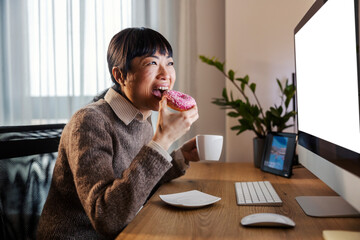 A hungry japanese businesswoman is eating doughnut at home office while looking at monitor.