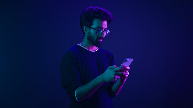 Indian man Arabian male model guy win mobile phone success victory smartphone achievement winning game cellphone internet betting online money prize at ultraviolet neon dark blue studio background
