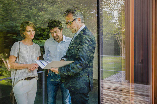 Paar mit Immobilienmakler schauen Vertragsunterlagen an einer neuen Immobilie,Blick durchs Fenster, Bayern, Deutschland