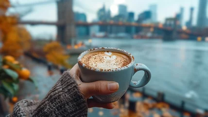  Close-up of a female hand holding a cup of coffee and Brooklyn Bridge is in the background, first-person photo, blurred background, travel image with well known destination © Loucine